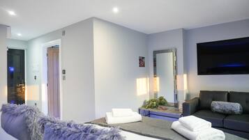 A modern hotel room with a grey sofa, a flat-screen TV, a large mirror, and neatly folded towels on a table.