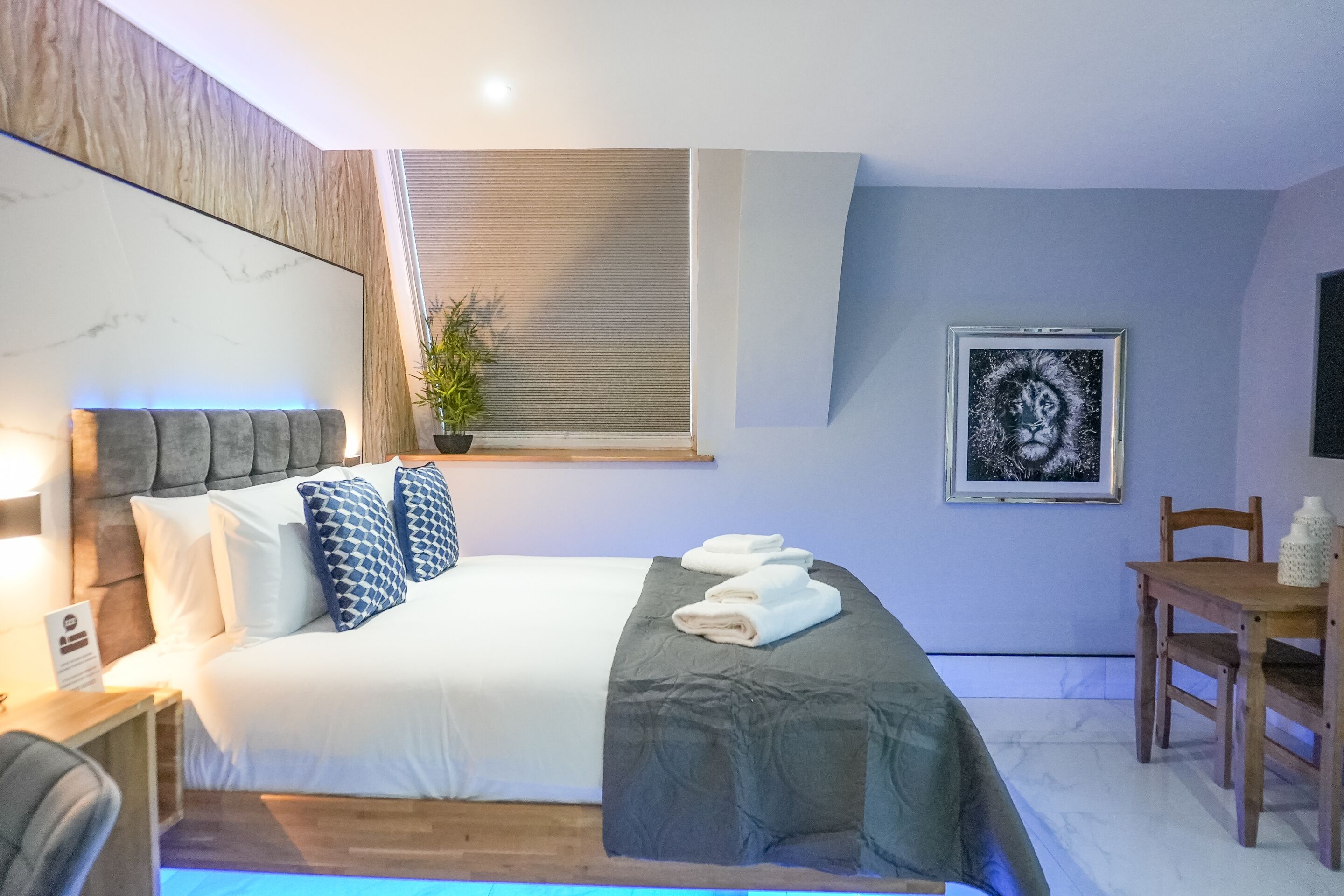 A modern hotel room with a large bed, a bedside table, a chair, a window with blinds, a framed artwork on the wall, and a small table with a bottle.