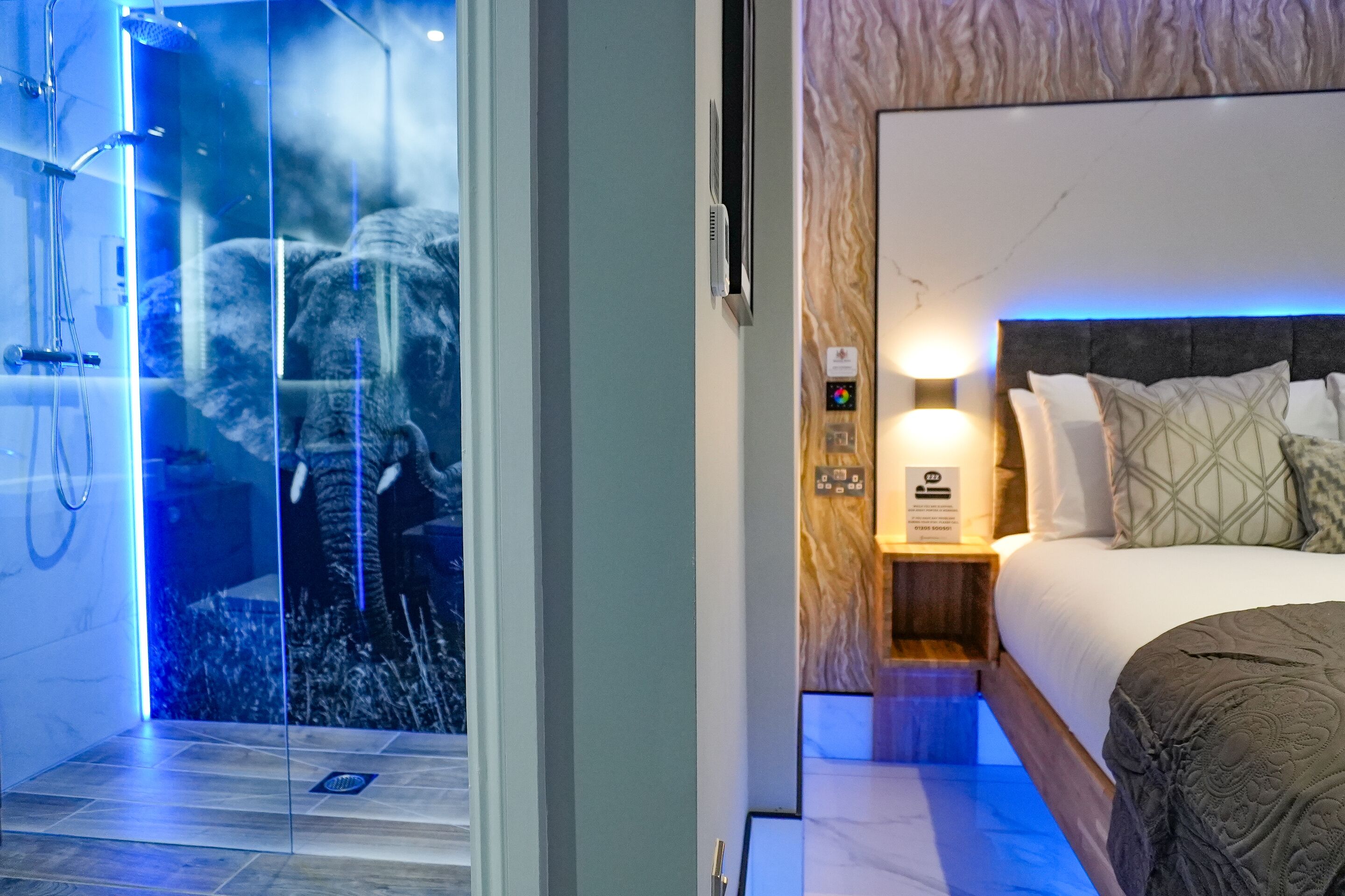 Quarto de hotel moderno com chuveiro em cabine de vidro, cama com encosto e mesa de cabeceira com abajur.