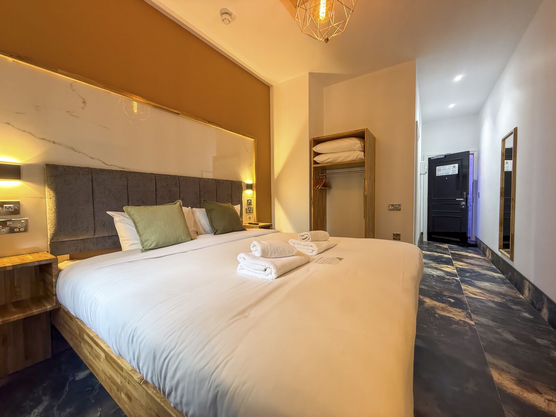 Ein Hotelzimmer mit einem großen Bett, einem Holzkopfende, einem Nachttisch, einer Wandlampe, einer eingebauten Ablage und einer Tür mit einem Spiegel.