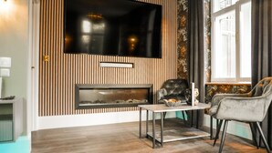 A modern living room with a flat-screen TV mounted above a fireplace, a comfortable armchair, and a small table with a bowl of fruit. - The Arundell Hotel (Boston)