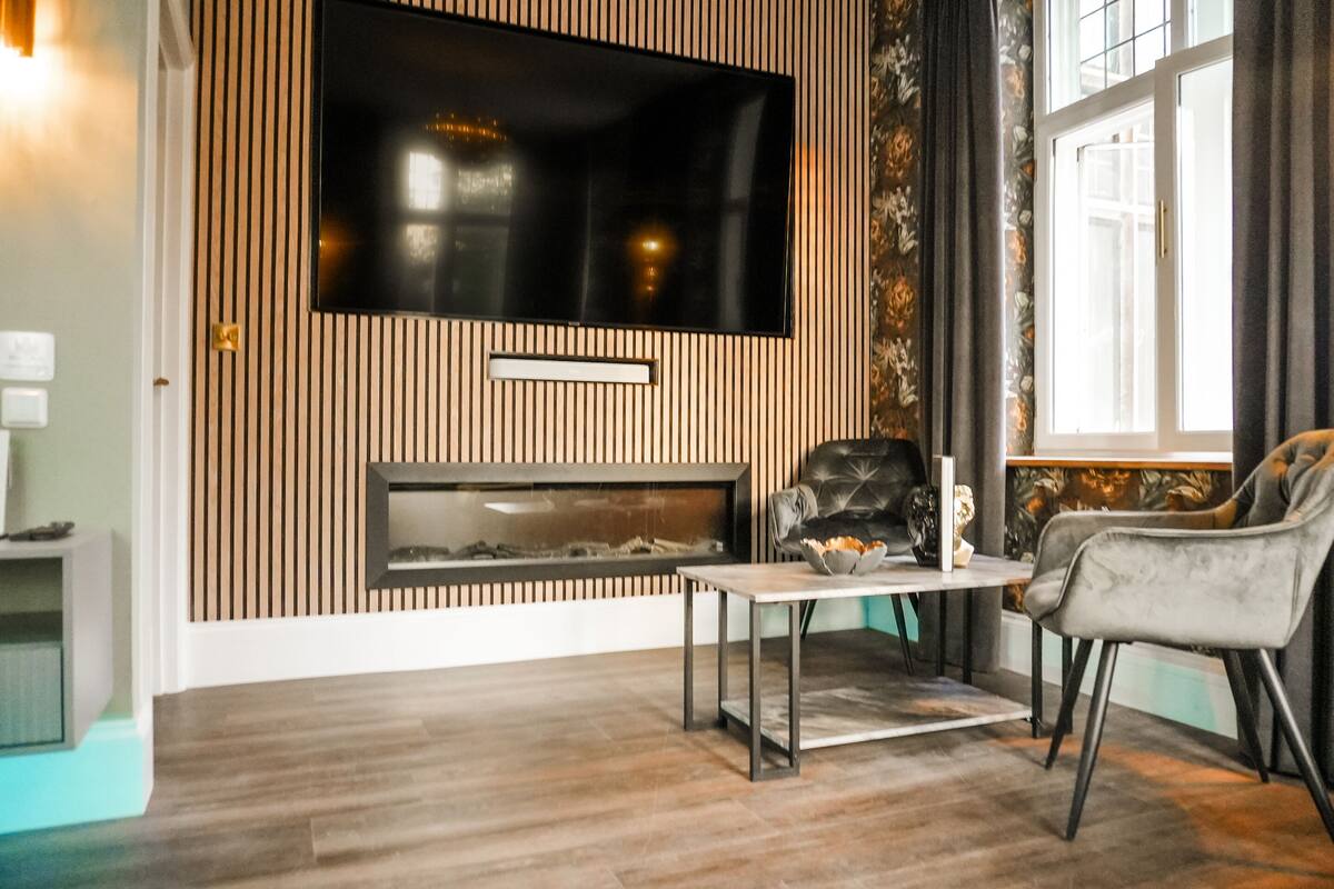 A modern living room with a flat-screen TV mounted above a fireplace, a comfortable armchair, and a small table with a bowl of fruit.