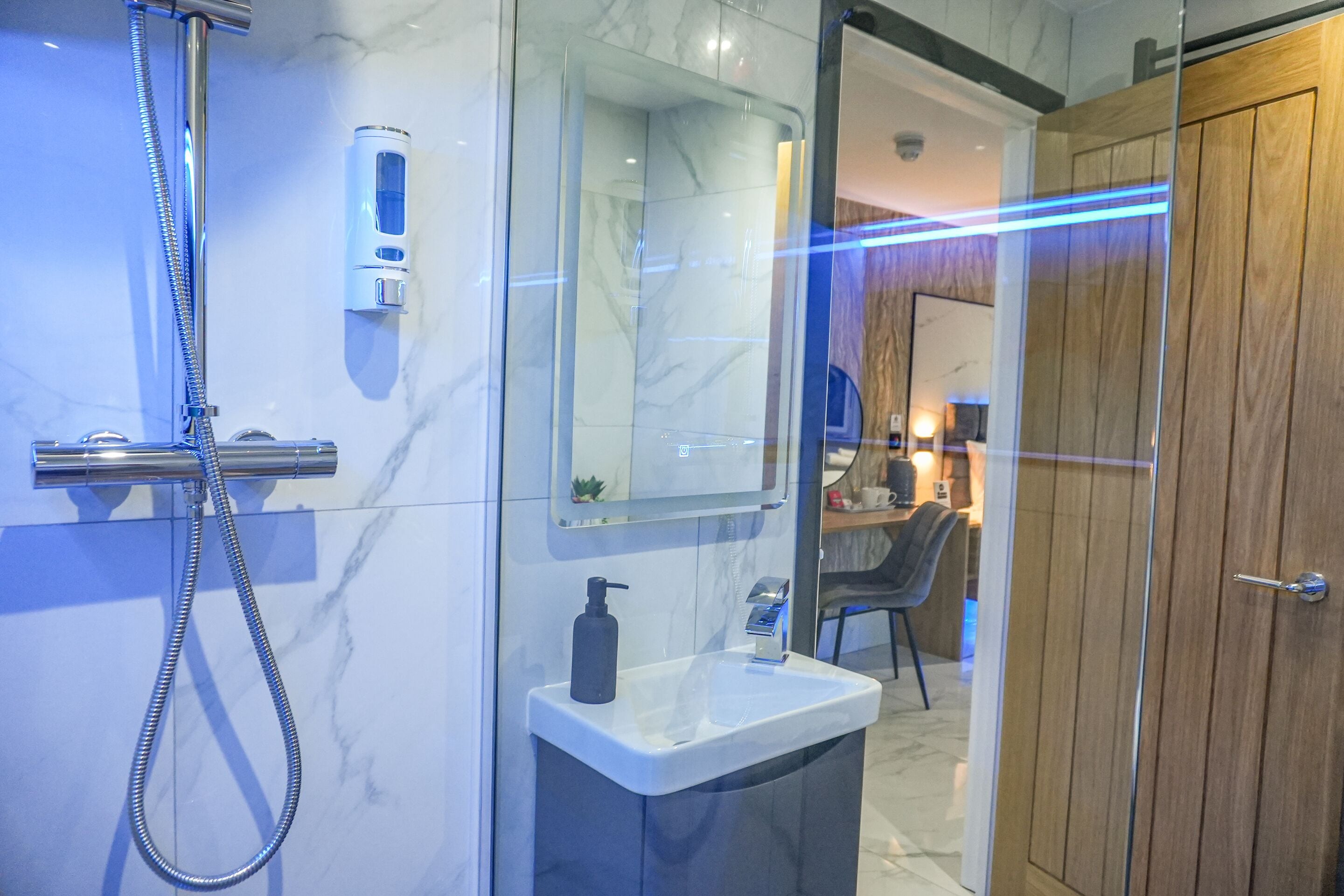 A modern bathroom with a glass shower enclosure, a marble-tiled wall, a wooden door, and a small sink with a soap dispenser.