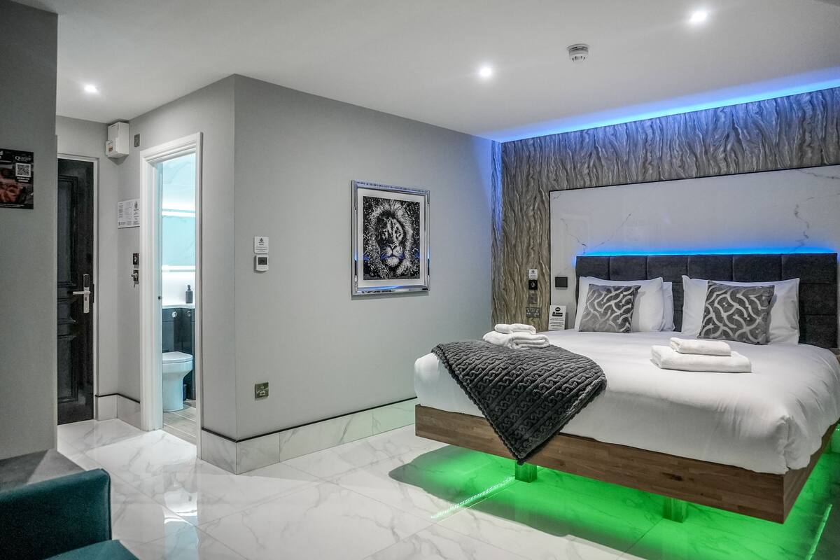A modern bedroom with a large bed, a headboard with blue lighting, a framed artwork on the wall, and a bathroom visible through an open door.
