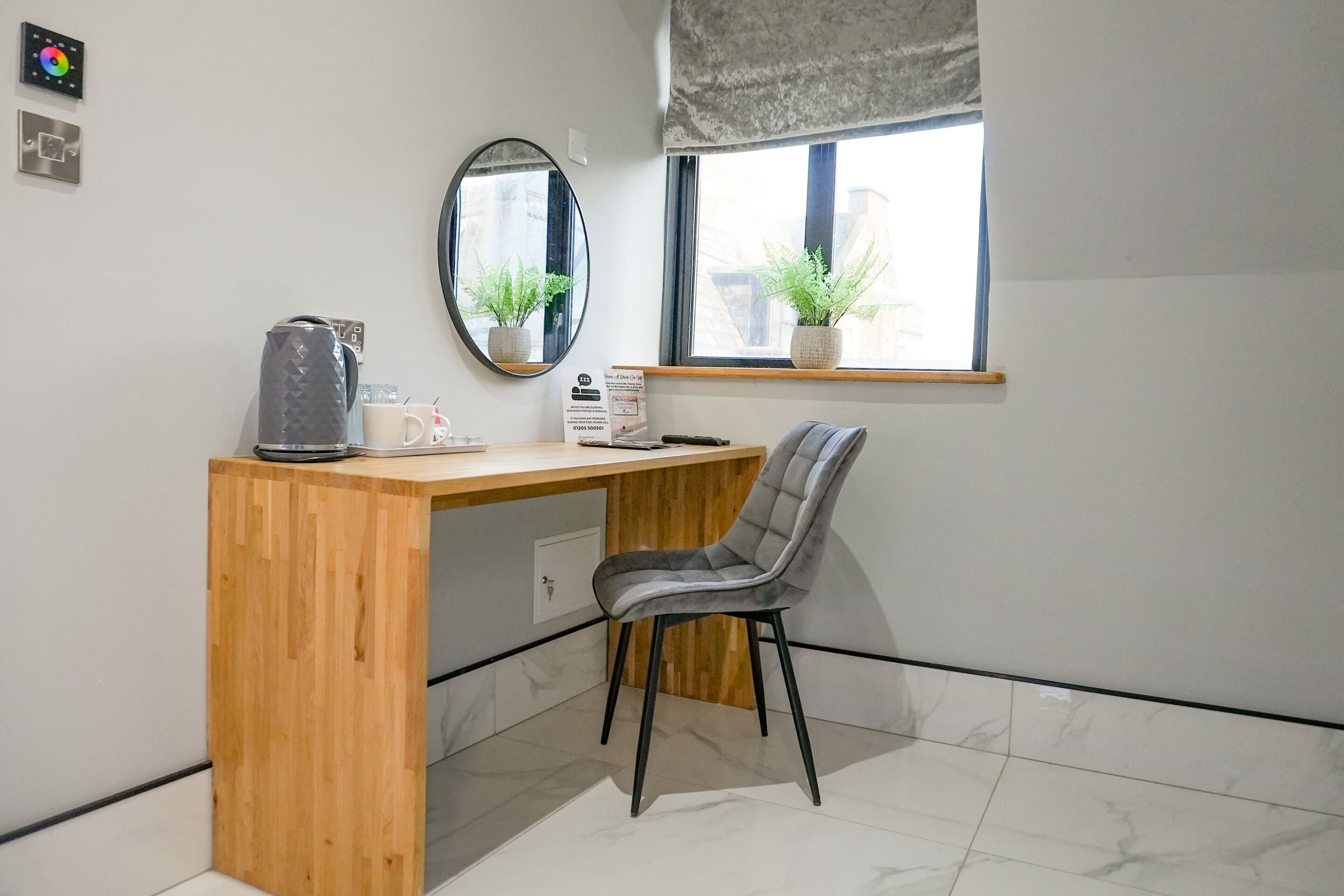 A small, modern room with a wooden desk, a grey chair, a mirror, and a window with plants.