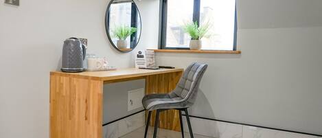 Una habitación pequeña y moderna con un escritorio de madera, una silla gris, un espejo y una ventana con plantas.