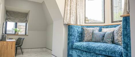 A room with a blue sofa, a wooden desk, and a chair. There is a window with curtains and a view of buildings outside.
