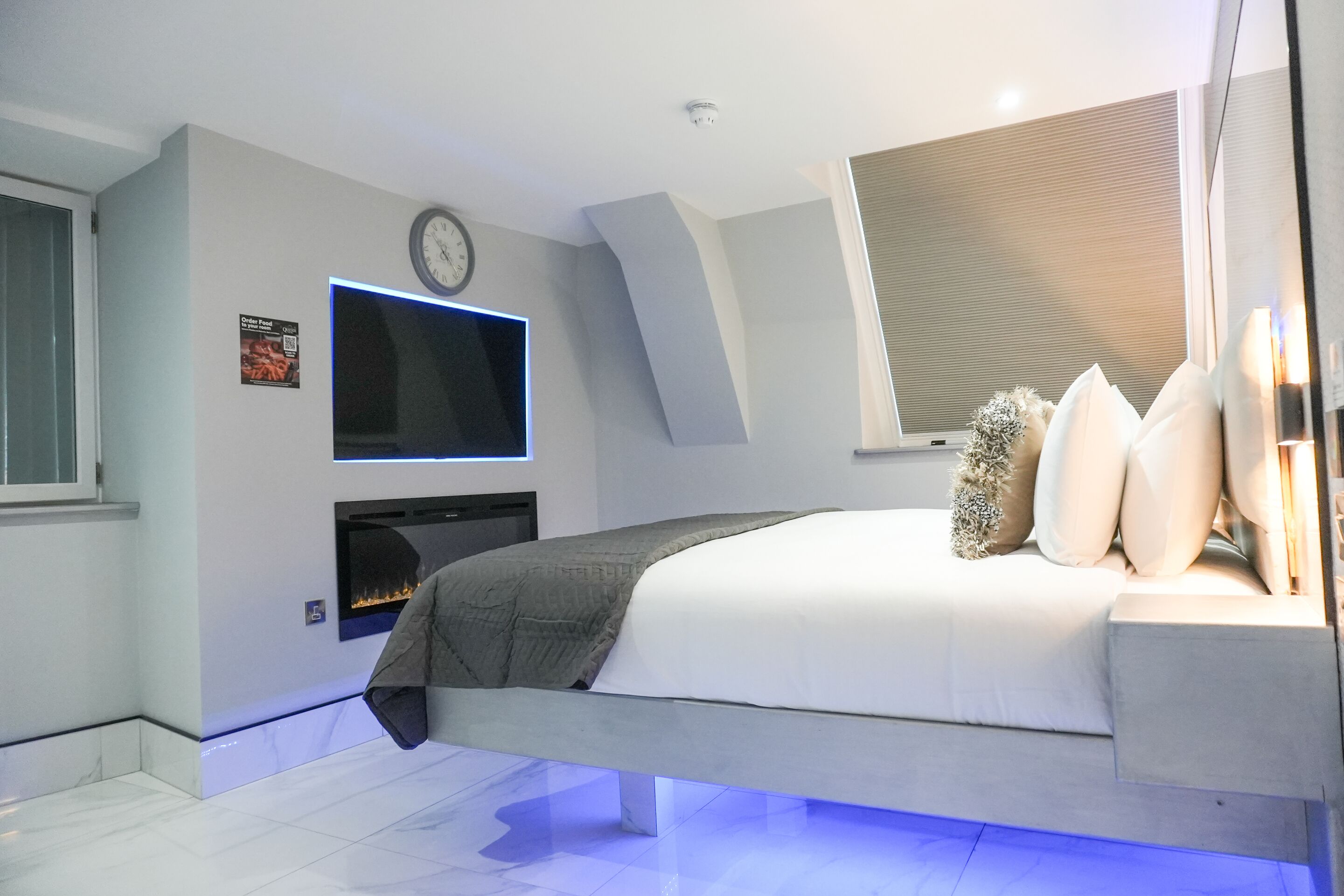 A modern bedroom with a bed, a flat-screen TV mounted on the wall, a fireplace, and a window with blinds.