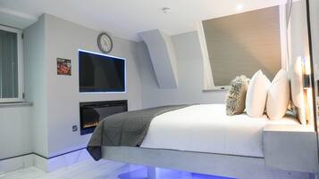 A modern bedroom with a bed, a flat-screen TV mounted on the wall, a fireplace, and a window with blinds.