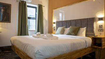 Una cama bien tendida con sábanas blancas y toallas enrolladas, un espejo grande encima y una ventana con cortinas verdes.