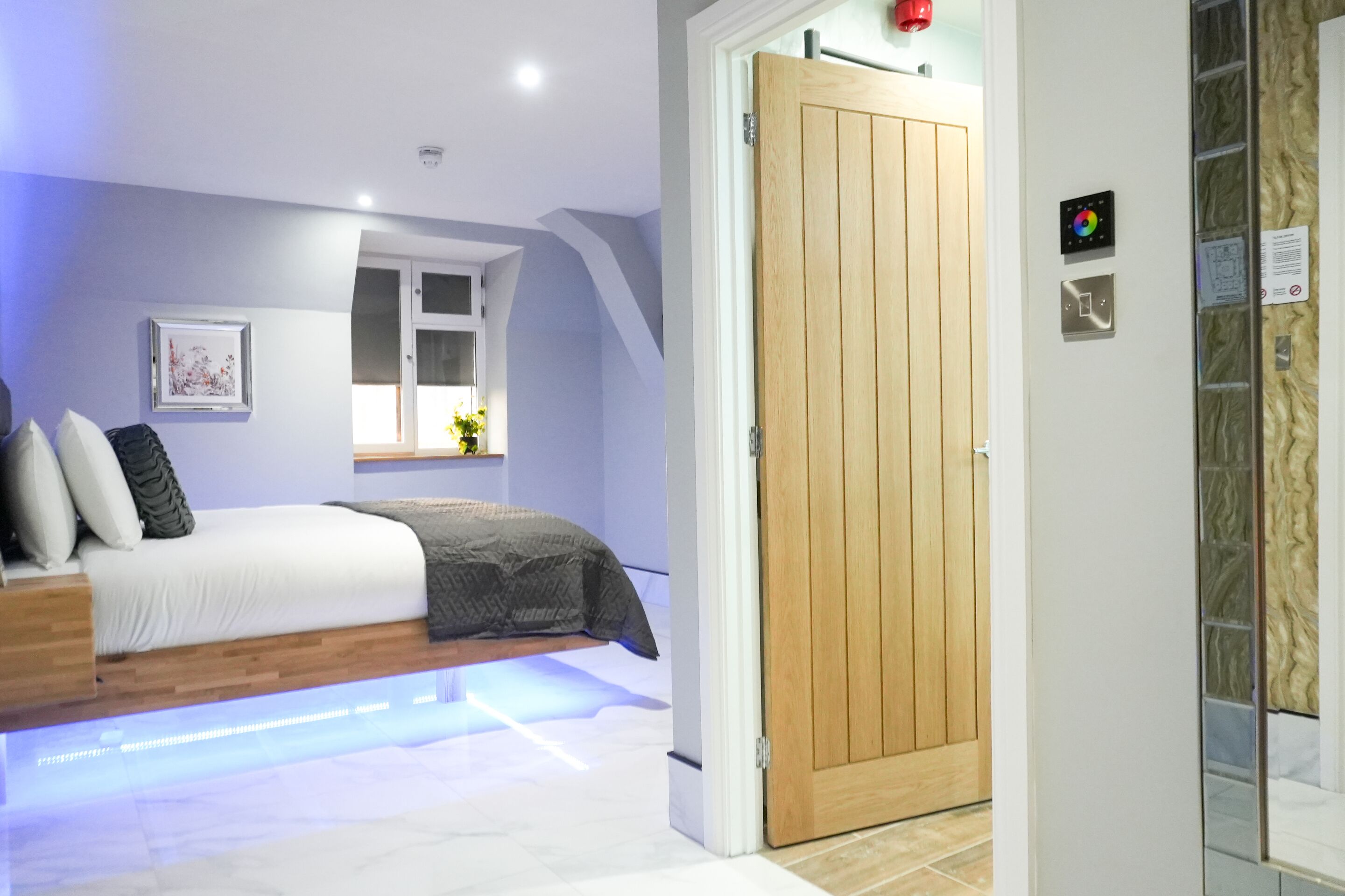 A bedroom with a bed, a window, a door, and a wall-mounted control panel.