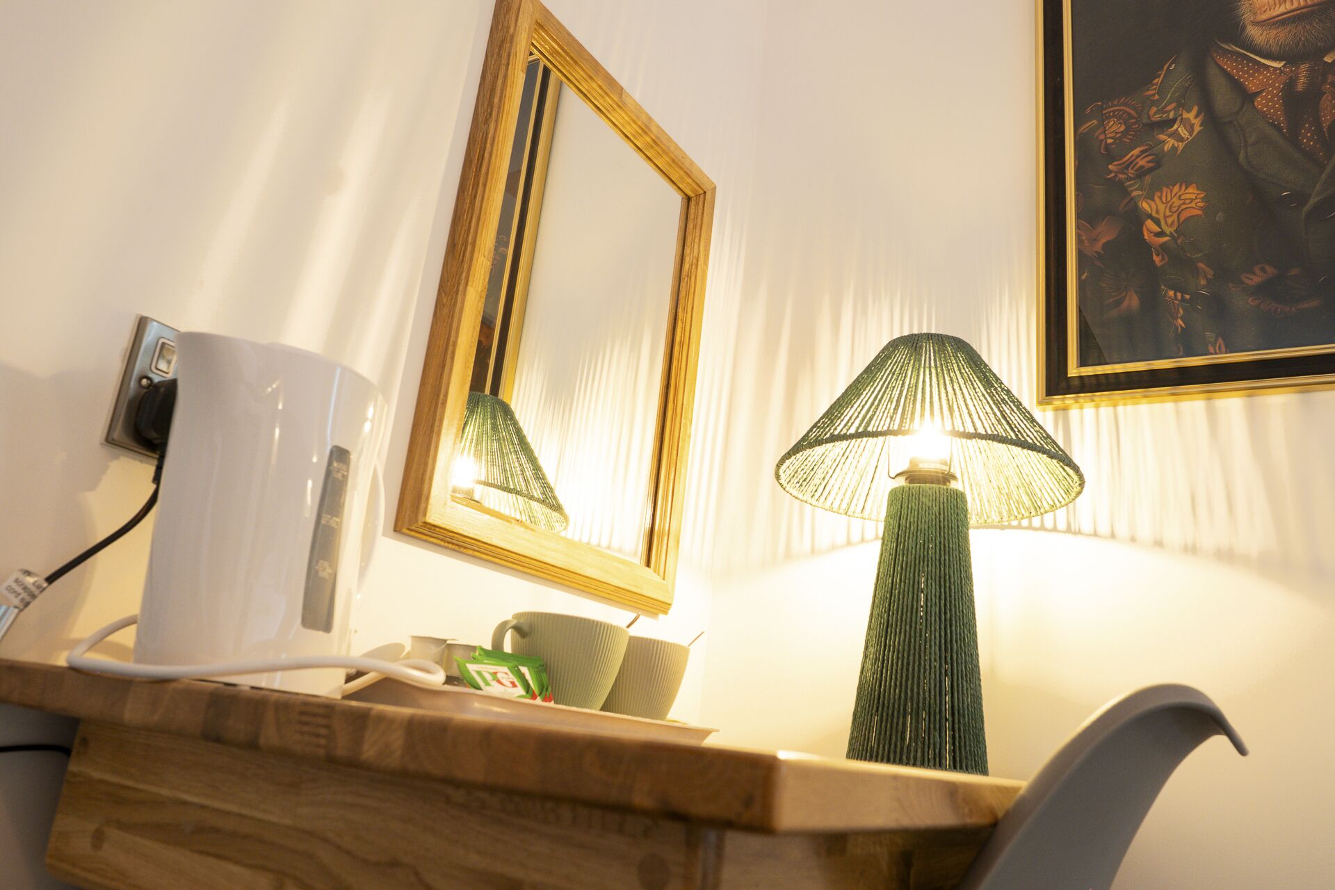A hotel room with a wooden desk, a green lamp, a white mug, and a framed picture on the wall.