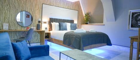 A modern hotel room with a large bed, a blue sofa, a small table, a round mirror, and a large window with blinds.