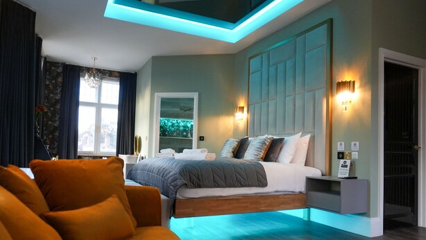 A modern bedroom with a bed, a sofa, a nightstand, and a wall-mounted light fixture. - The Arundell Hotel (Boston)