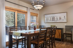 A dining area with a wooden table, chairs, a vase with flowers, and a painting of deer on the wall.