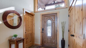 A wooden door with a glass panel, a round mirror, a welcome sign on a wooden table, and a doormat with a bear design.