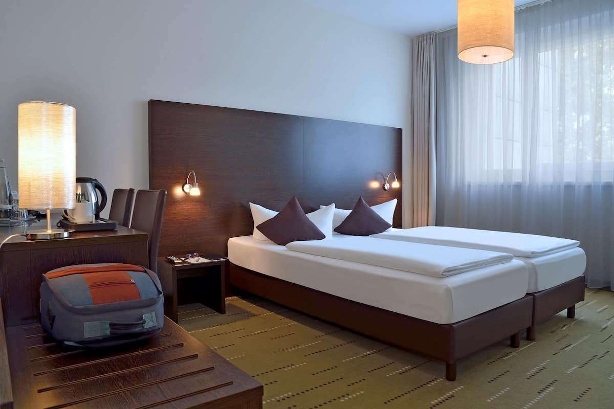 A hotel room with a bed, bedside tables, a lamp, a suitcase, and a view of the outdoors.