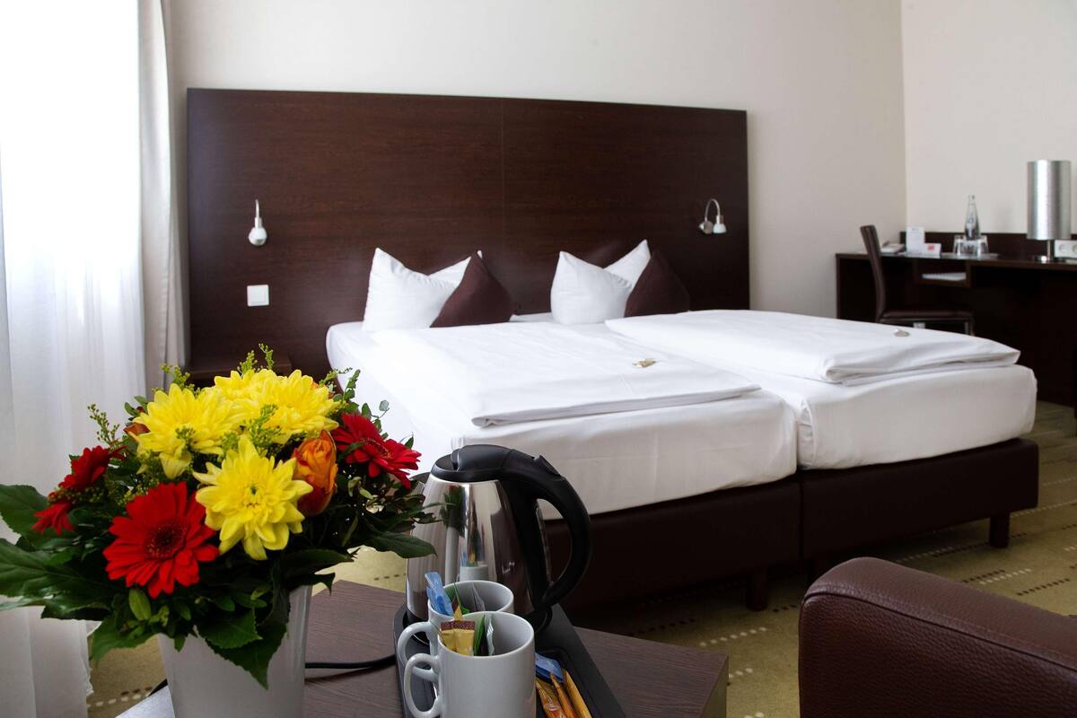 A hotel room with a double bed, a desk with a chair, a coffee maker, and a bouquet of flowers.