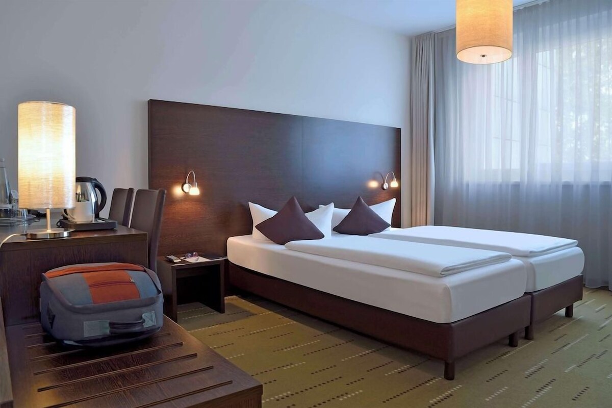 Ein Hotelzimmer mit einem großen Bett, einem Schreibtisch mit Lampe, einem Sessel und einem Koffer.