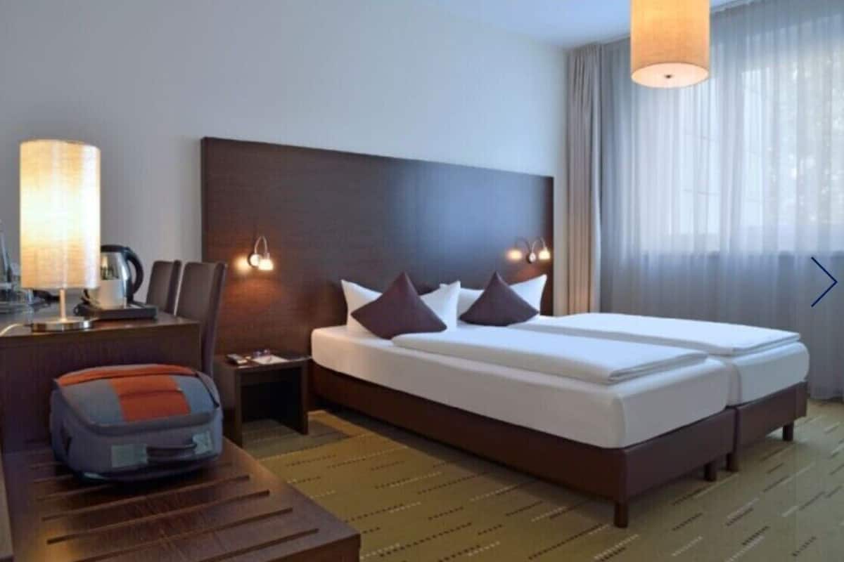 A hotel room with a large bed, two bedside tables, a desk with a lamp, and a suitcase on the floor.