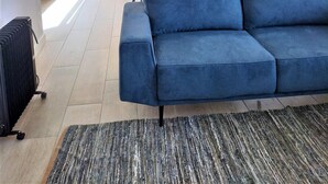 A modern living room with a blue sofa, a dining table with chairs, and a large window providing natural light. - T2 Requinte A C e Vista Mar Wifi Garagem (Loulé)