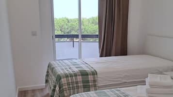 A hotel room with two beds, a large window with curtains, a ceiling fan, and a view of greenery outside.