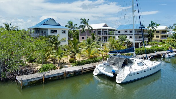 Un port de plaisance où un voilier est amarré, entouré d’une végétation tropicale et bordé d’une rangée de maisons aux toits bleus.