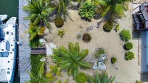 Luchtbeeld van een jachthaven met een aangemeerd schip, omgeven door palmbomen en een zandstrand met een houten terras en ligbedden.