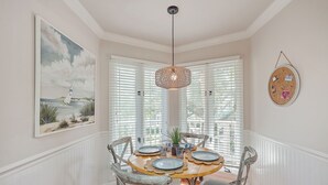 A dining area with a wooden table, four chairs, a hanging light fixture, and a painting of a sailboat on the wall. - Port O' Call E203 - Ocean View Perfection! (Isle of Palms)