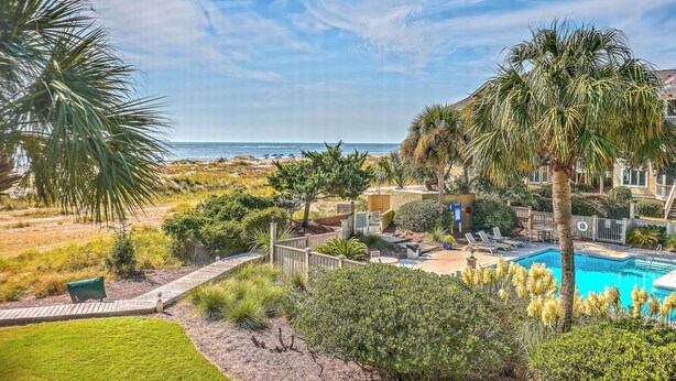 A pool area with a view of the ocean, surrounded by palm trees and a well-maintained lawn. - Port O' Call E203 - Ocean View Perfection! (Isle of Palms)