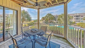 A covered outdoor area with a table and chairs, a ceiling fan, and a view of a swimming pool and buildings in the background. - Port O' Call E203 - Ocean View Perfection! (Isle of Palms)