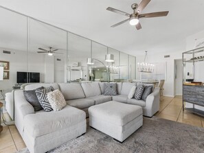 A modern living room with a sectional sofa, a ceiling fan, and a large mirror reflecting the space.