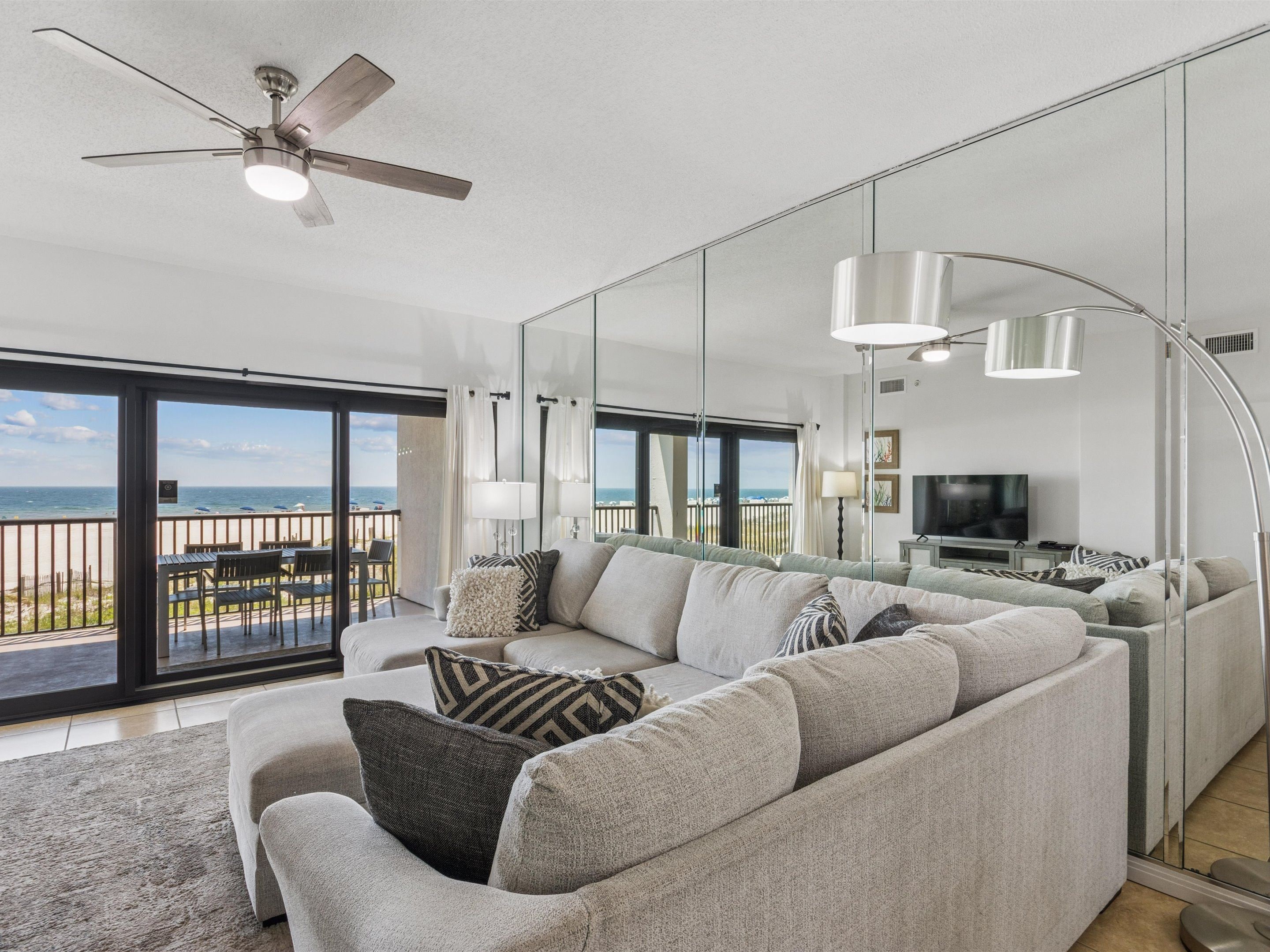 Een moderne woonkamer met een grote bank, een plafondventilator en uitzicht op het strand door grote glazen deuren.