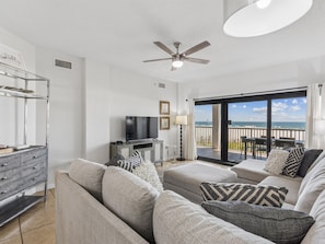 Een moderne woonkamer met een flatscreen televisie, grijze banken en een balkon met uitzicht op zee.
