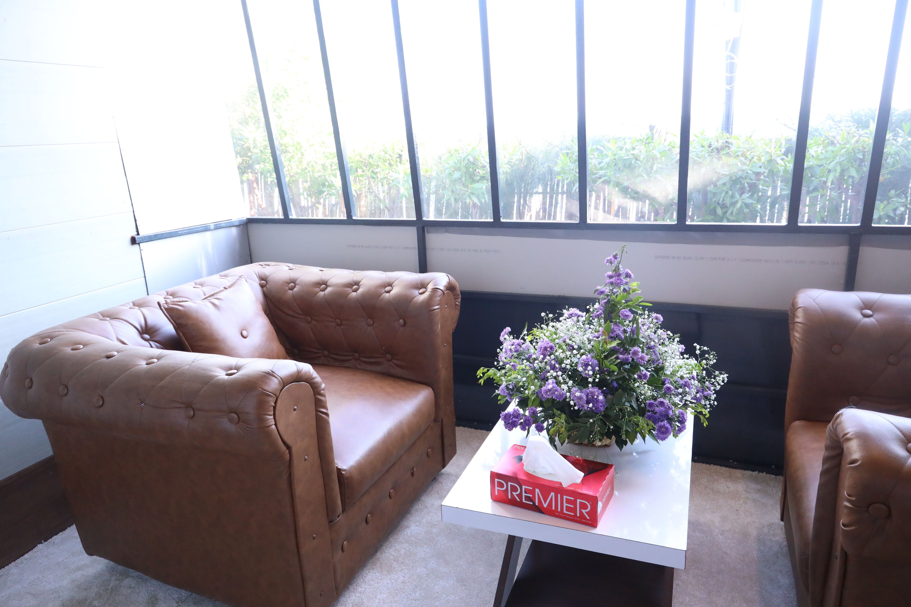 Una habitación con dos sillones de cuero marrón, una mesa blanca con una caja roja etiquetada “PREMIER” y un jarrón con flores moradas.