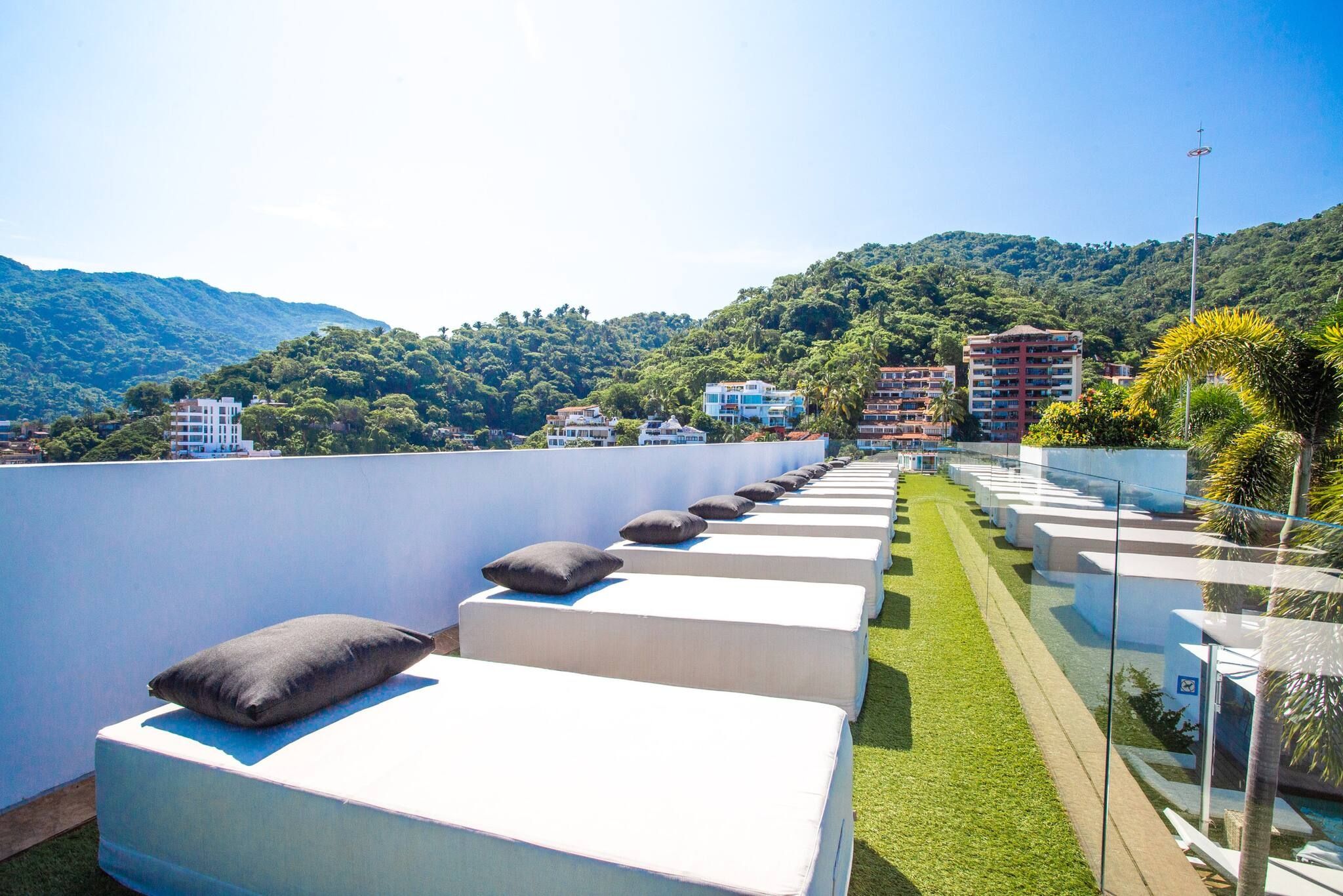 A rooftop terrace with white cushioned seating and a glass railing overlooking a mountainous landscape.