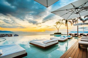 An infinity pool with lounge chairs and umbrellas, overlooking a sunset over the sea.
