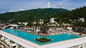 Uma grande piscina ao ar livre com várias áreas de descanso e palmeiras, cercada por edifícios e um cenário montanhoso.