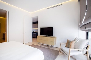 A modern hotel room with a bed, a TV, a sofa, and a kitchenette in the background.