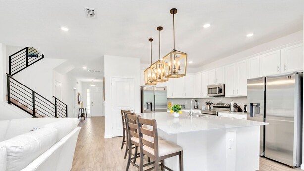 A modern kitchen with white cabinets, a large island, stainless steel appliances, and a staircase with black railings.