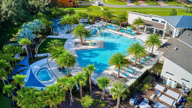 Vista aérea de un resort con una gran piscina rodeada de palmeras, un área de estar techada y un edificio residencial al fondo.