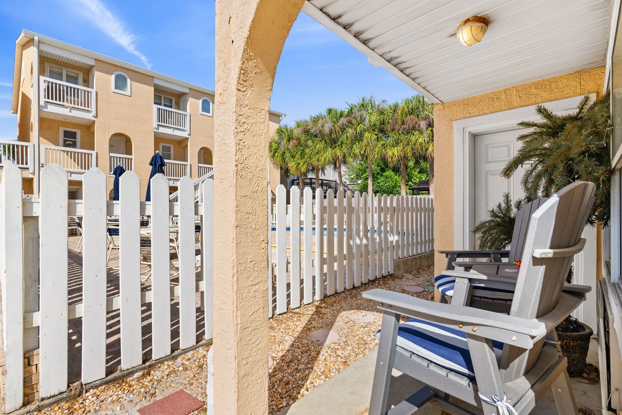 A covered outdoor seating area with a white picket fence, a pool in the background, and a building with balconies.
