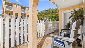 A covered outdoor seating area with a white picket fence, a pool in the background, and a building with balconies.