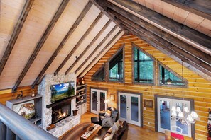 A log cabin interior with a stone fireplace, wooden beams, and large windows.