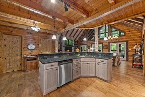 A spacious kitchen with a central island, wooden cabinetry, and a stone fireplace. - Mountaintown Creek Lodge – Creekside Cabin with Hot Tub, Game Room & Fishing (Ellijay)