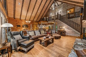 A spacious log cabin interior with a high ceiling, a dining area, and a staircase leading to an upper level. - Mountaintown Creek Lodge – Creekside Cabin with Hot Tub, Game Room & Fishing (Ellijay)