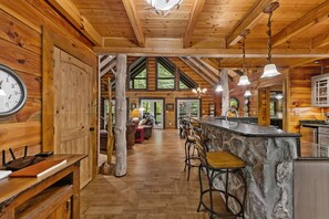 A log cabin interior with a central bar area, wooden floors, and a large clock on the wall. - Mountaintown Creek Lodge – Creekside Cabin with Hot Tub, Game Room & Fishing (Ellijay)