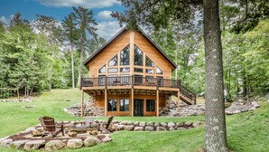 A two-story log cabin with a balcony, surrounded by a stone retaining wall and a grassy yard. - NEW! Gorgeous Northwoods Home on the Eagle River Chain of Lakes (Eagle River)