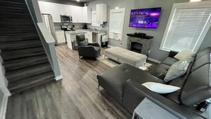 A modern living room with a gray sofa, a fireplace, and a kitchen area with white cabinets and a flat-screen TV. - Practically Perfect Place in Every Way (Pensacola)