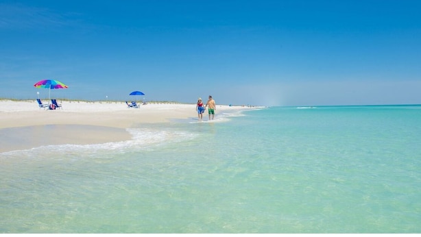 A beach with clear turquoise water, a few people walking, and colorful umbrellas on the sand. - Practically Perfect Place in Every Way (Pensacola)